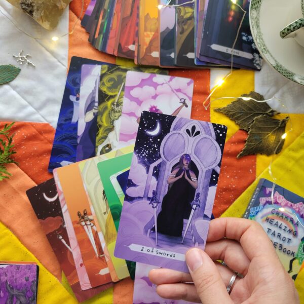 A few of the swords cards from Chakra Healing Tarot deck held by a hand with a colourful background: a quilted Tarot cloth, surrounded by flatlay arrangement of Chakra Healing Tarot cards, the guidebook, some leaf-shaped charms, a crystal, a teacup and saucer, and small fairy lights.
