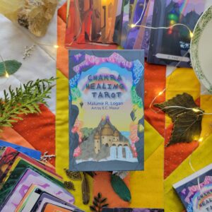 Chakra Healing Tarot deck in a sturdy and colourful box rests on a quilted Tarot cloth, surrounded by flatlay arrangement of Chakra Healing Tarot cards, the guidebook, some leaf-shaped charms, a crystal, a teacup and saucer, and small fairy lights.