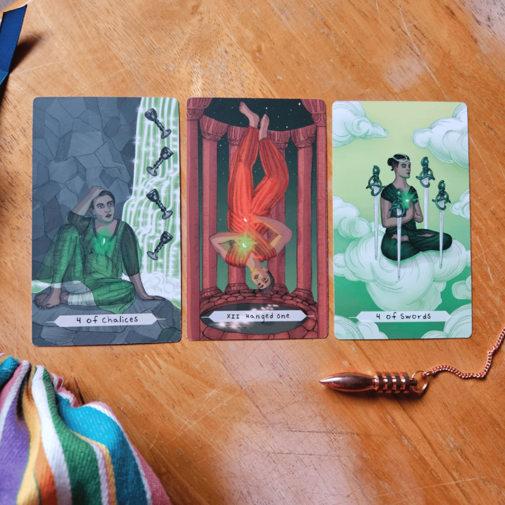 Three Tarot cards on a wooden table: 4 of Chalices, Hanged One, 4 of Swords. There is also a copper pendulum and a colourful cloth bag on the table.