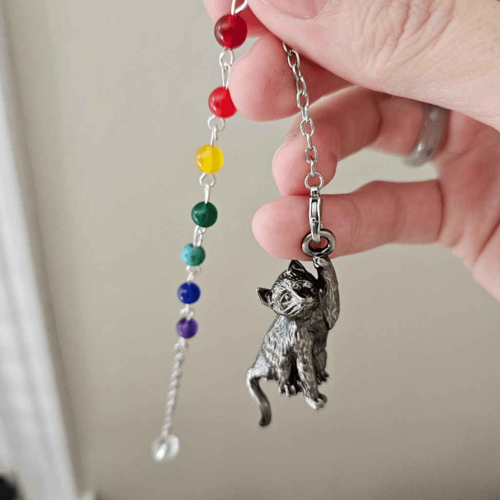 A person's hand holds a pendulum featuring a cat-shaped swinging pointer and seven stones (red, orange-red, yellow, green, turquoise, dark blue, purple) with a clear bead at the end of chain.