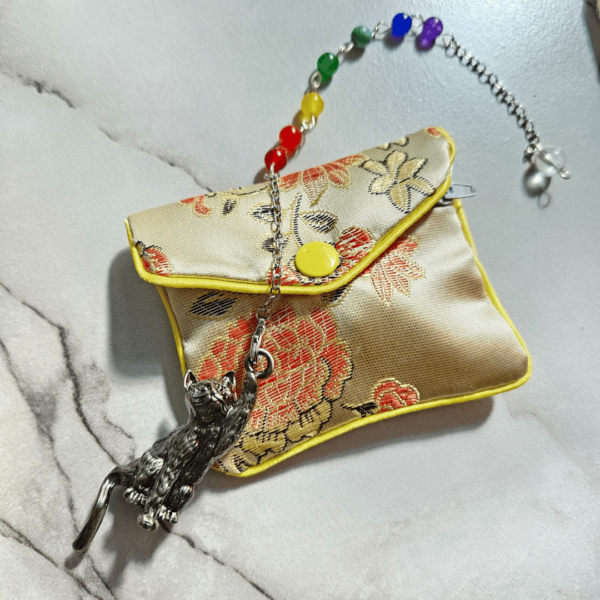A pendulum with a swinging cat-shaped pointer rests on a pale gold brocade pouch on a marble-like surface. The pendulum chain has seven small stones in the chain, starting from the cat going upward toward a clear bead at the end: deep red, orange-red, yellow, green, turquoise, deep blue, purple.