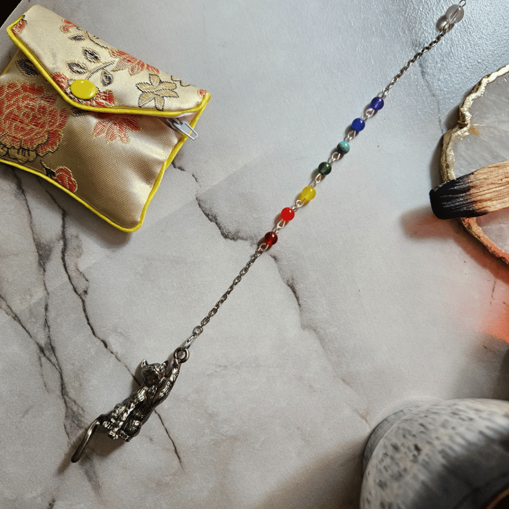 A pendulum featuring a cat-shaped swinging pointer and seven stones (red, orange-red, yellow, green, turquoise, dark blue, purple) rests on a marble-like surface. There is a clear bead at the end of chain. A pale gold brocade pouch is also in the image. There is a glimpse of a coffee mug, white and gold coaster and a used stick of palo santo to the sides/corner of the image.