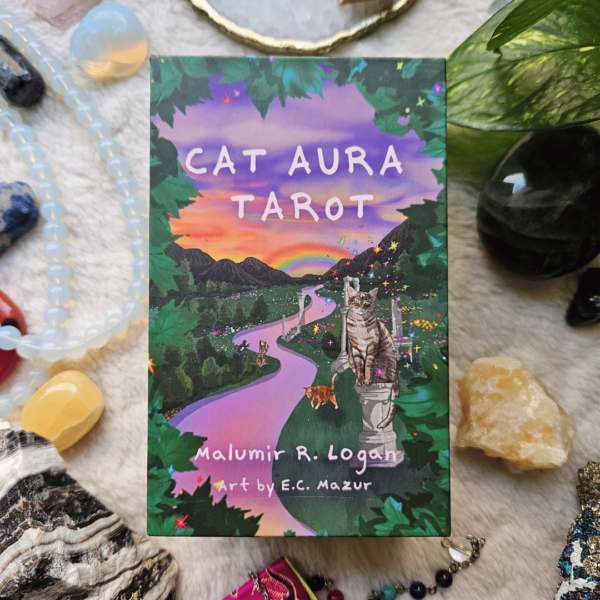 Cat Aura Tarot deck box rests on a soft creamy surface, surrounded by crystals, a plant, a journal and pen.