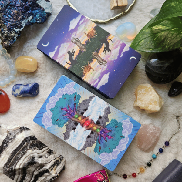 Flatlay image of Chakra Healing Tarot and Cat Aura Tarot decks resting side by side diagonally toward the upper left on a soft creamy surface with crystals, a plant and a cat-shaped pendulum also in the scene.