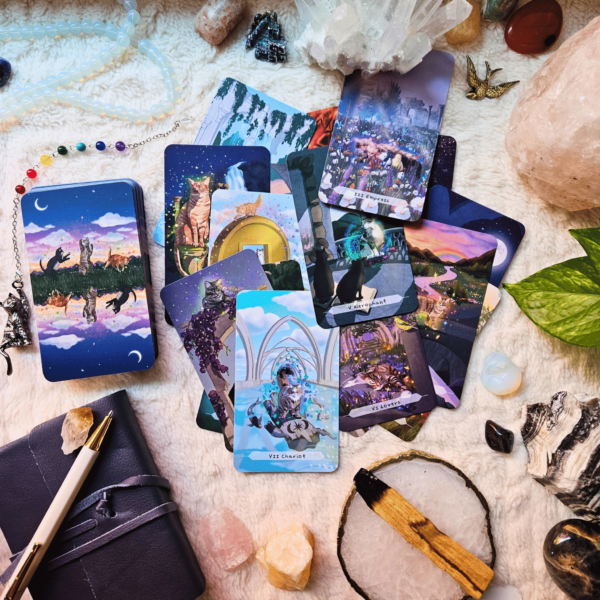 Flatlay image of Cat Aura Tarot deck on a fluffy cream surface, surrounded by crystals, a plant, a teacup and saucer, palo santo, a necklace, and a journal and pen.