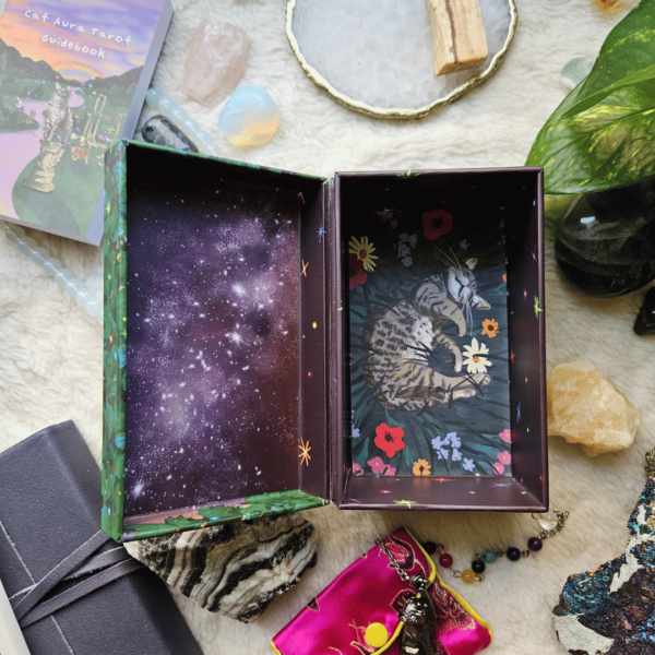 Cat Aura Tarot deck box rests open on a soft creamy surface, surrounded by crystals, a plant, a journal and pen. The inside of the box top depicts a starry sky at night and the bottom depicts a sleeping cat, curled up in soft grass and flowers.