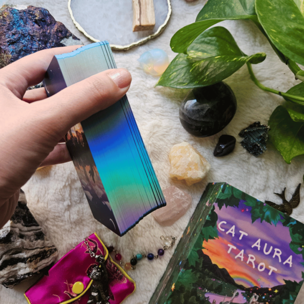 Cat Aura Tarot deck is edged with light blue rainbow holographic edging. A hand holds this deck to show the edging, above a surface covered with a fluffy creamy covering and a flatlay scene with crystals, a plant, and Cat Aura Tarot Guidebook.