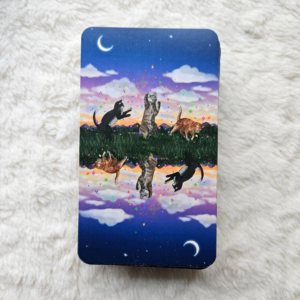 Cat Aura Tarot card backs depict a fully reversible image of three cats playing with colourful sparks under a late dusk sky that hosts a few clouds, stars and a crescent moon.