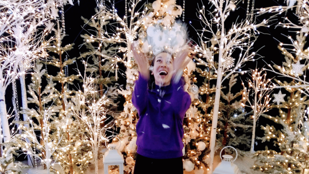 A person in a purple top and black pants throws fake snow into the air jubilantly above themselves in the foreground. In the background are festive tree and plant shaped decorations with soft white lights, illuminated against a dark backdrop.