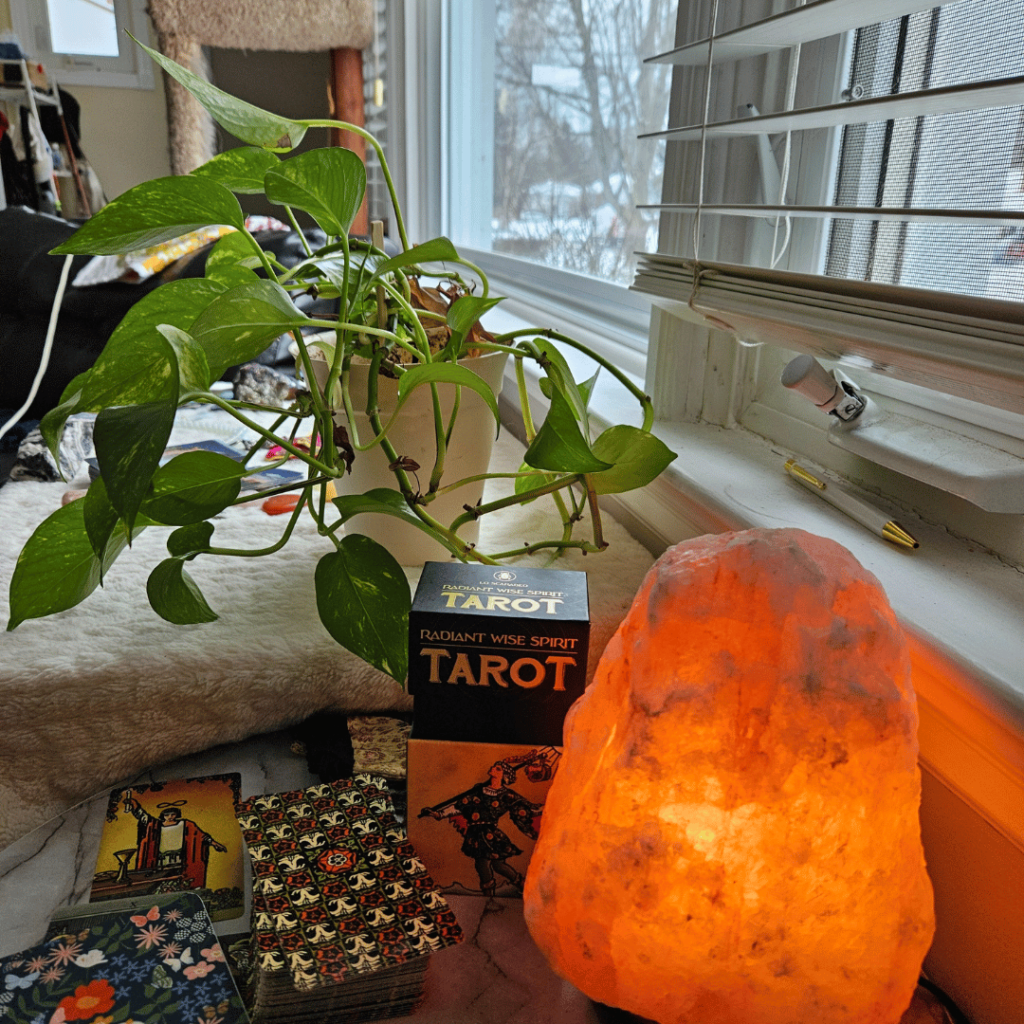 A view of a small table hosting a salt lamp, Tarot cards, notebook, and another table with a plant on it, all in front of a window.