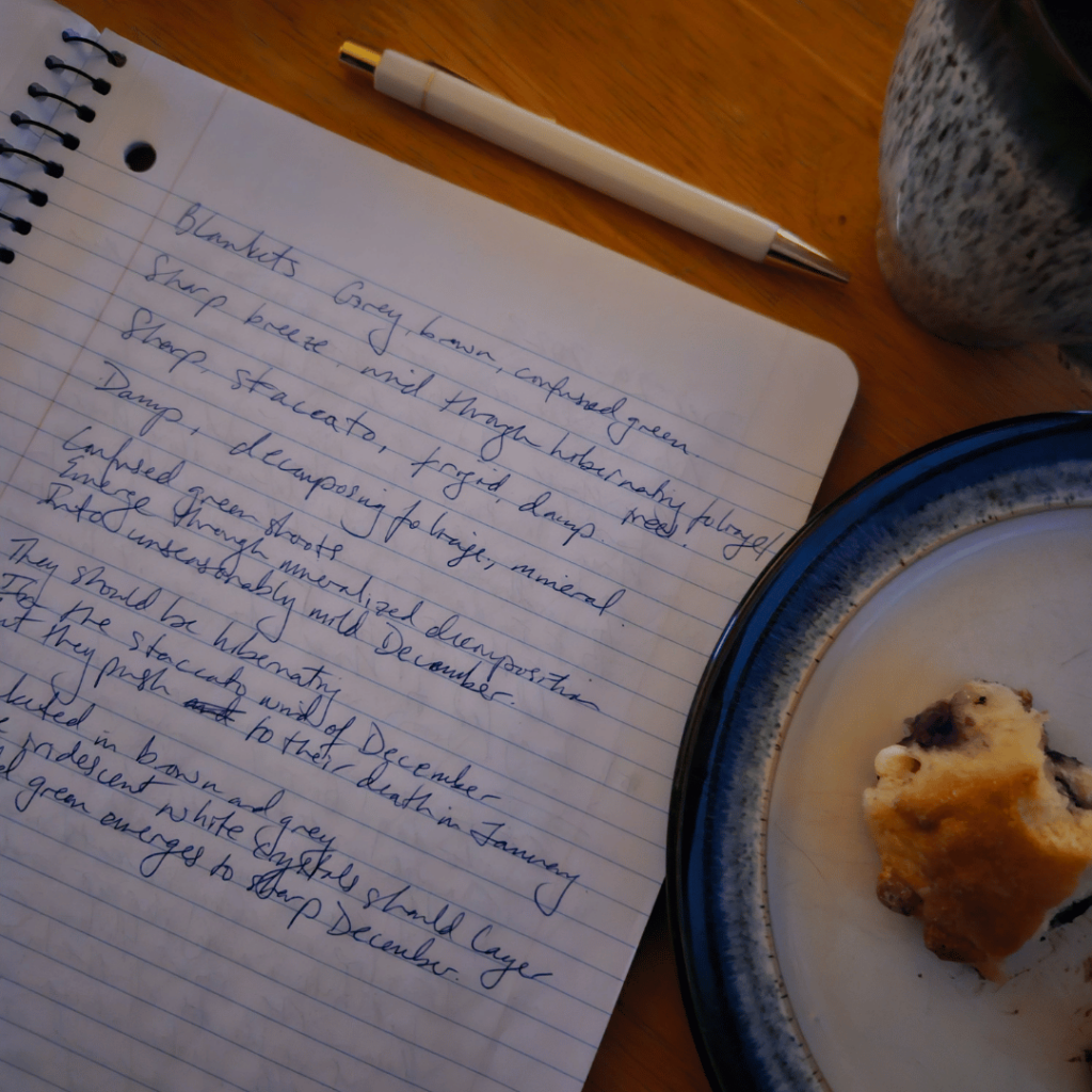A notebook with scrawled handwriting in blue ink rests on a wooden surface, a light-coloured pen with metal details resting above the notebook. A mug and plate with a partially eaten berry scone are also on the table.